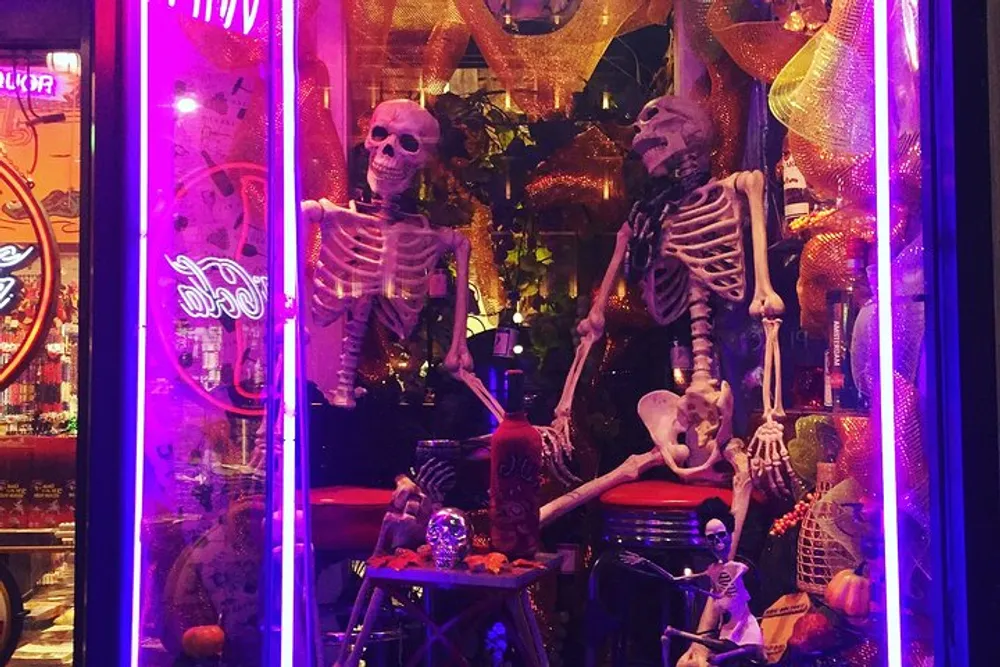 The image shows a vibrant Halloween-themed window display with two life-size skeletons amidst festive decorations illuminated by a purple neon light