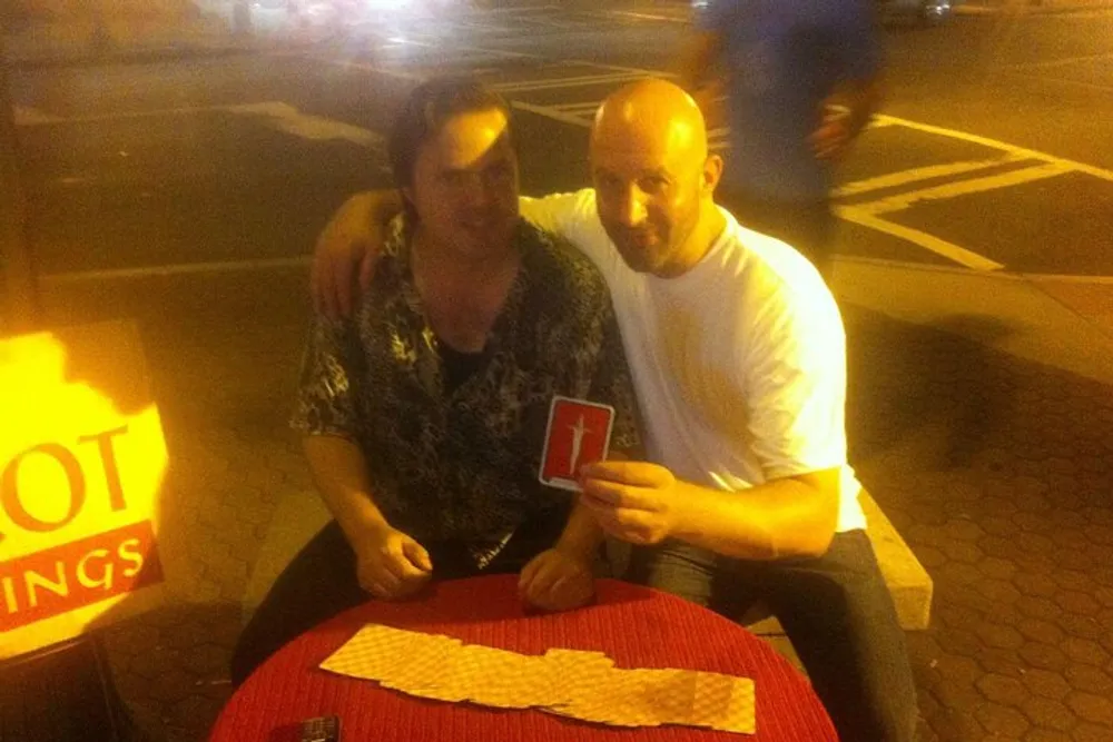Two people are posing at a table with one person holding up a red playing card against a blurred night-time streetscape background