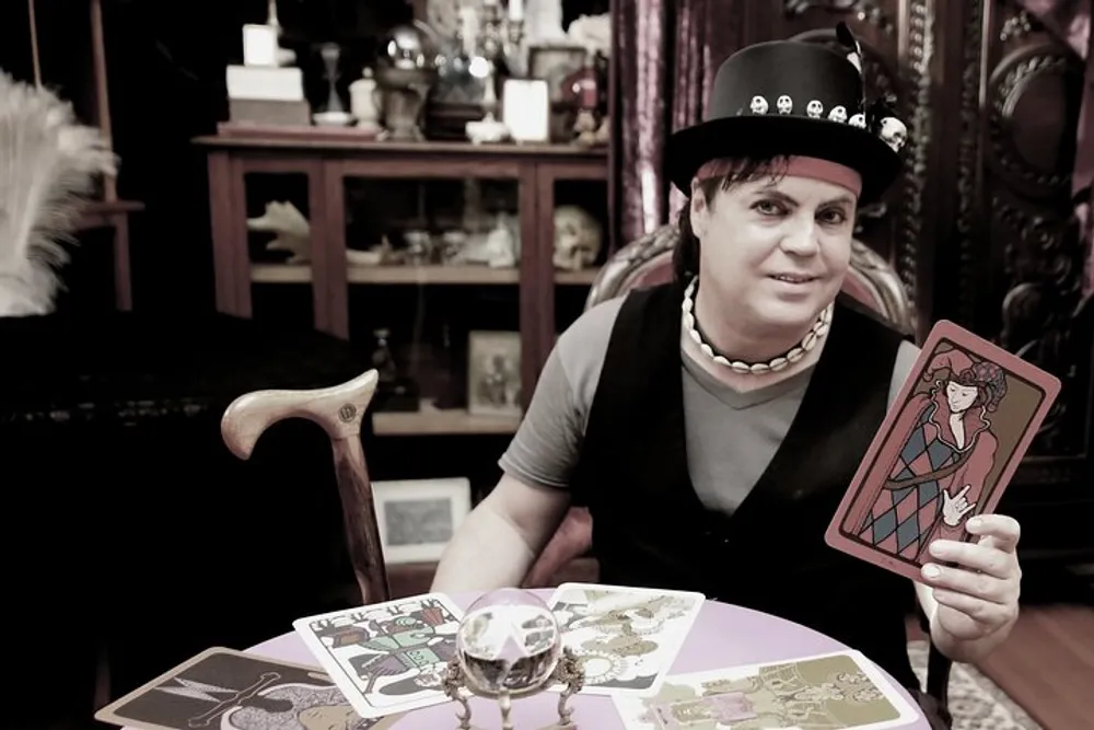 A person is sitting at a table with tarot cards spread out holding a card towards the camera with a crystal ball in front of them in a room filled with various decorative items