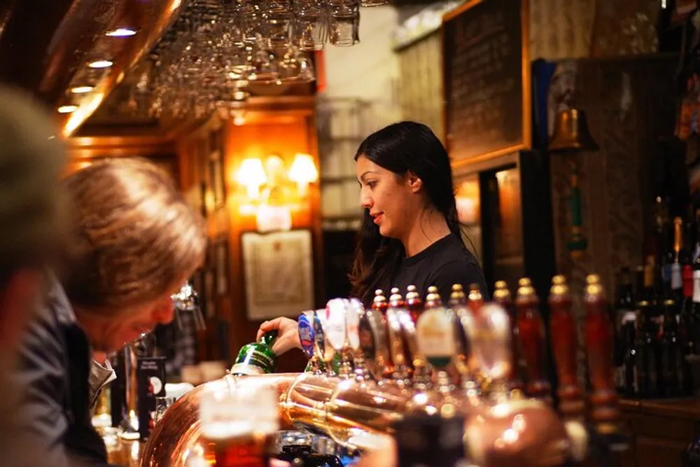 A bartender is pouring a drink at a cozy warmly-lit pub with customers at the bar