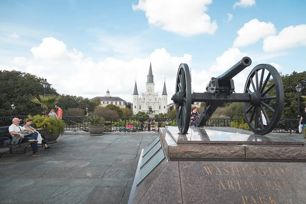 The image shows a historical cannon on display at Washington Artillery Park with people relaxing on benches and the iconic spires of the St Louis Cathedral in the background under a partly cloudy sky