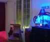 The image depicts a dimly lit room with eclectic decor featuring colored lights a high-back armchair a dresser with decorative objects and a lamp casting a blue glow near a window with green light coming through