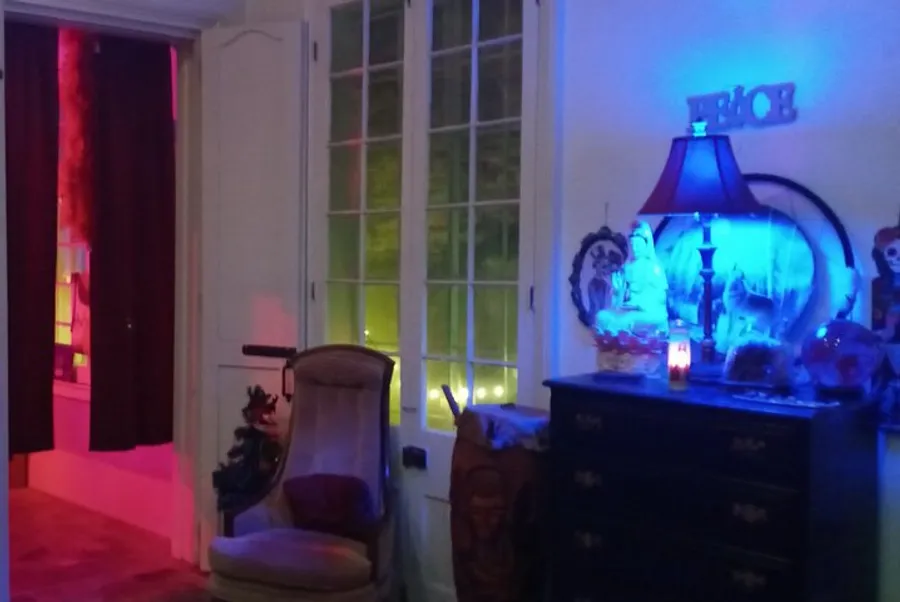 The image depicts a dimly lit room with eclectic decor, featuring colored lights, a high-back armchair, a dresser with decorative objects, and a lamp casting a blue glow near a window with green light coming through.