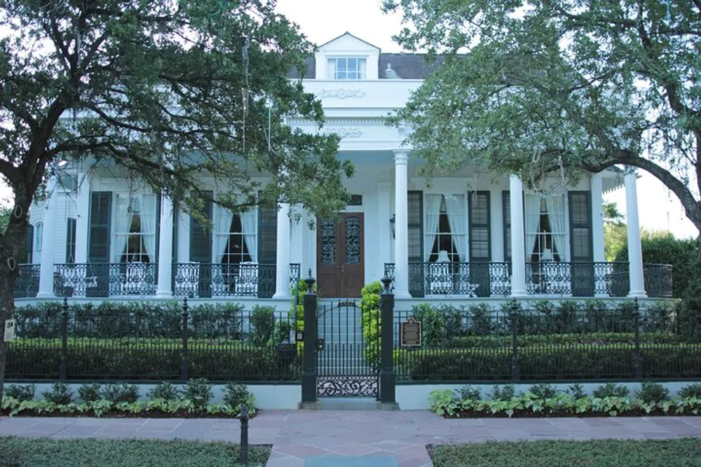The image shows a grand white house with a symmetrical facade framed by ornate black ironwork and lush greenery exuding antebellum charm