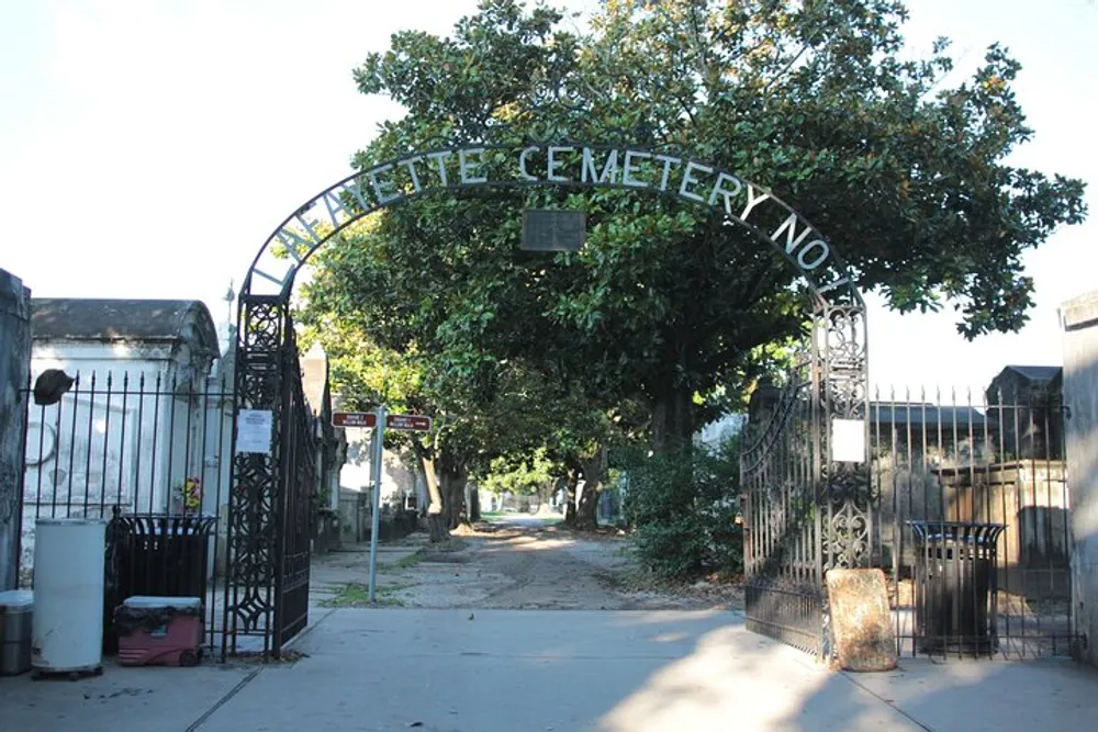 The image shows the entrance to Lafayette Cemetery No 1 with ornate metal gates opening to a tree-lined path through the historic cemetery