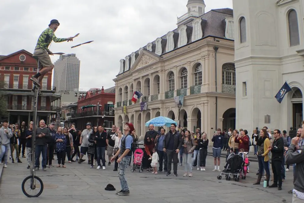 A street performer on a tall unicycle juggles clubs in front of an attentive crowd in a city square with historic buildings in the background
