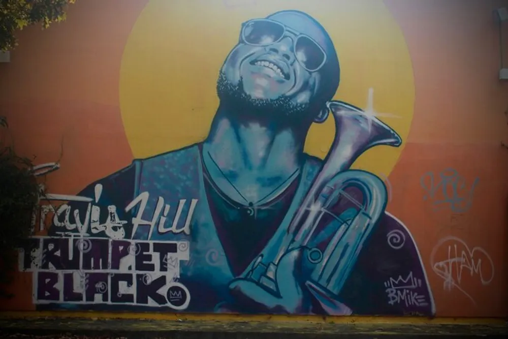 This image features a vibrant mural of a smiling person with sunglasses holding a trumpet with stylistic text and signatures around it