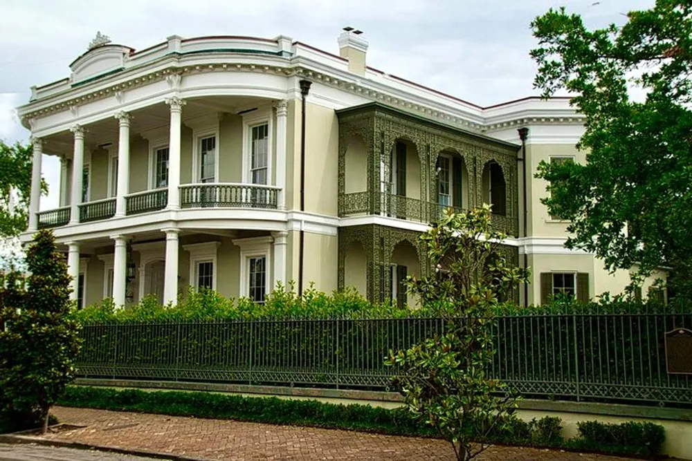 The image shows a grand two-story mansion with ornate ironwork on the balconies surrounded by a wrought iron fence and lush greenery