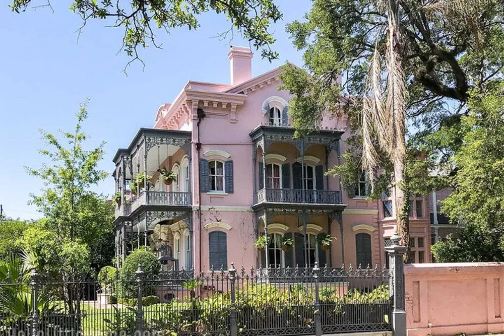 The image shows a pink Victorian mansion with a black wrought-iron balcony situated behind an ornate metal fence and surrounded by trees and greenery