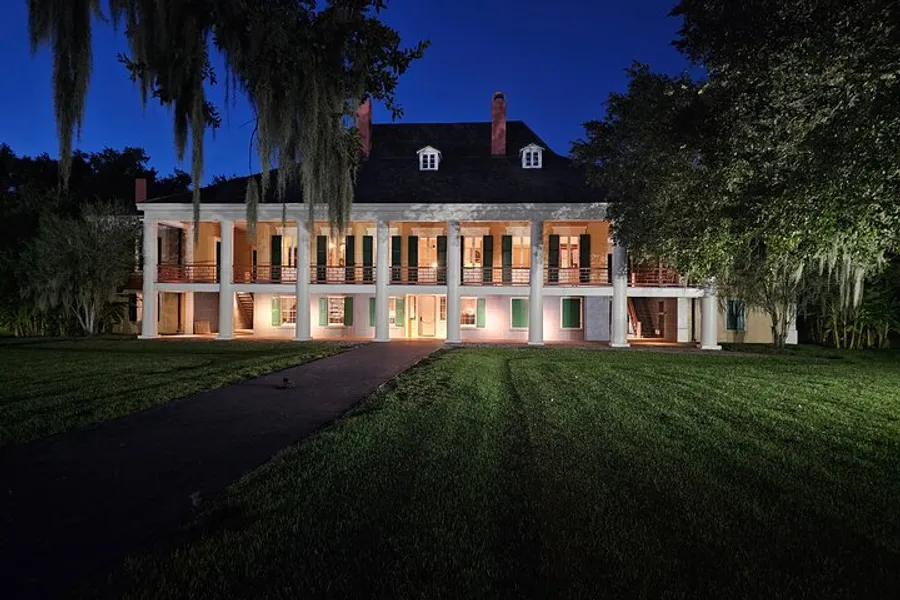 The image depicts a historic two-story plantation-style house with a symmetrical facade, illuminated by warm lights, at twilight with a lush green lawn in the foreground and draped Spanish moss adding an eerie ambiance.