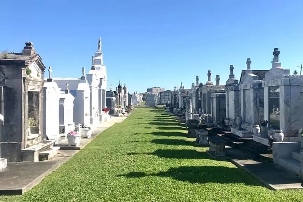 The image shows a sunny day at a cemetery with rows of above-ground tombs and statues characteristic of a historic or possibly New Orleans-style graveyard under a clear blue sky