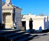 The image shows a sunny day at a cemetery with rows of above-ground tombs and statues characteristic of a historic or possibly New Orleans-style graveyard under a clear blue sky