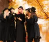 A group of five people dressed in black with hats some looking curious or whispering to each other are standing amid a golden autumnal scene with one person holding a book