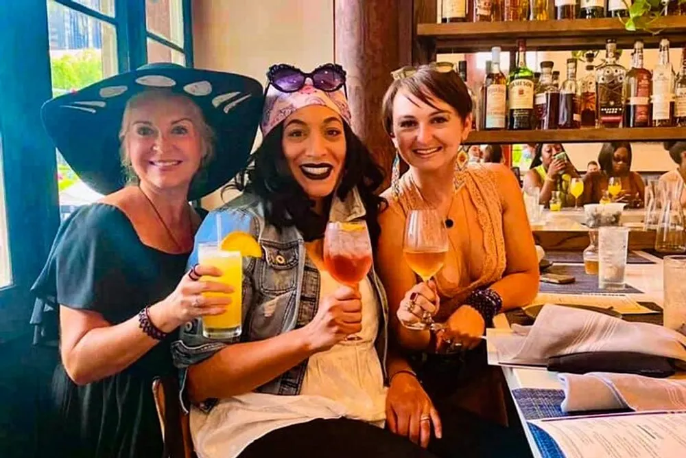 Three women are smiling and toasting with drinks at a bar or restaurant