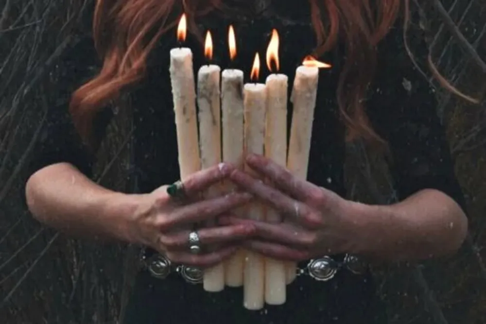 The image shows a person with red hair holding several lit white candles in their hands