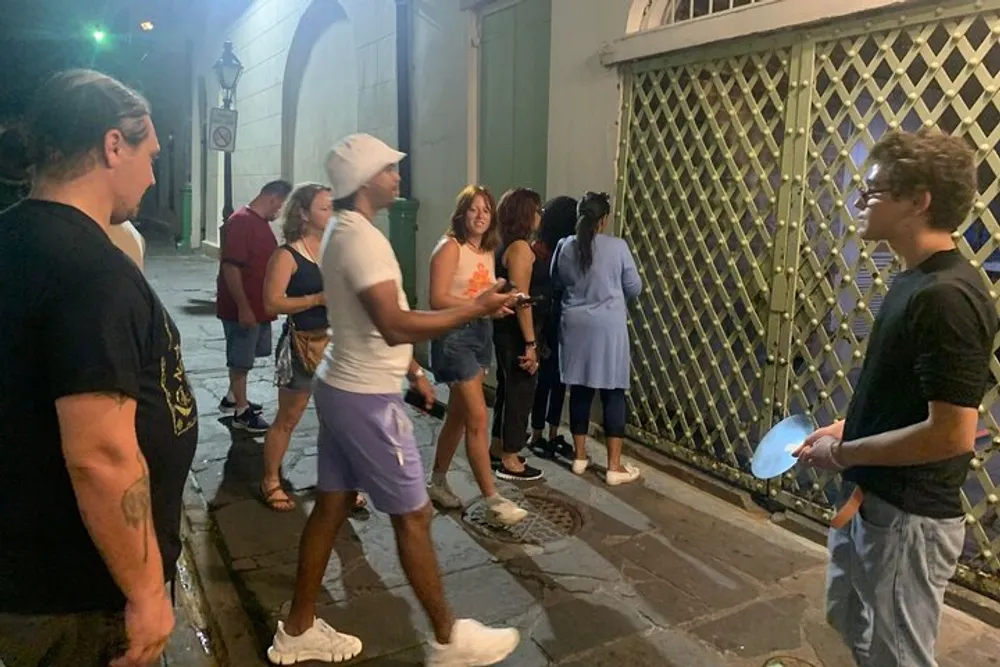 The image shows a group of people interacting on a city street at night with a man in white headwear gesturing mid-conversation and another holding a blue fan suggesting a social or nightlife scene