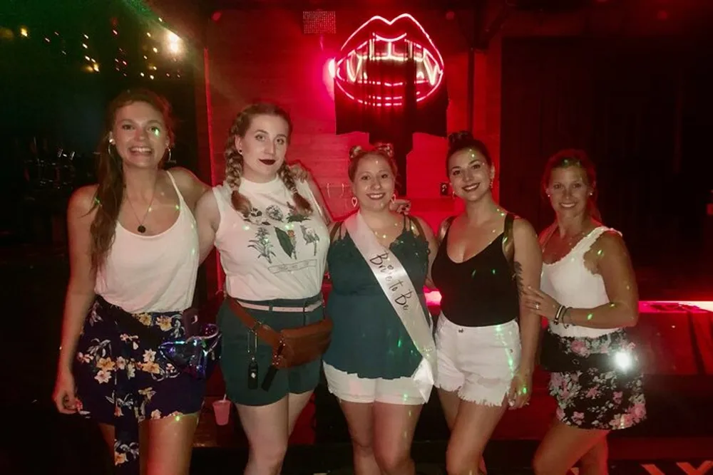 A group of five smiling women posing for a photo with one wearing a sash that reads Bride To Be indicating a bachelorette party or bridal celebration in front of a neon lip sign