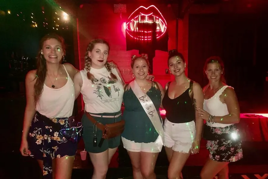 A group of five smiling women posing for a photo, with one wearing a sash that reads Bride To Be, indicating a bachelorette party or bridal celebration, in front of a neon lip sign.