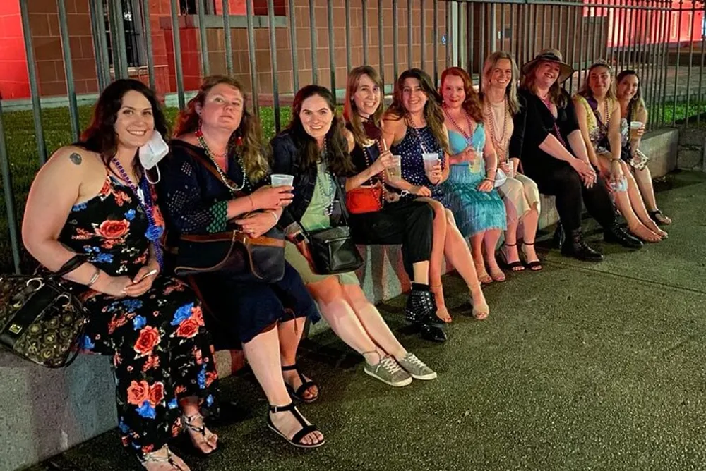 A group of people is sitting together on a low wall at night smiling and posing for a photo some holding beverages and they are all dressed in casual evening attire