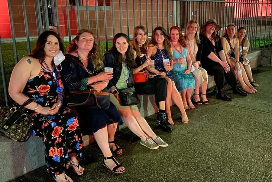 A group of people is sitting together on a low wall at night, smiling and posing for a photo, some holding beverages, and they are all dressed in casual evening attire.