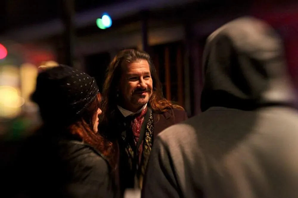 A man with long hair and a scarf smiles while engaging in a conversation with two people whose backs are to the camera against a blurred nighttime background