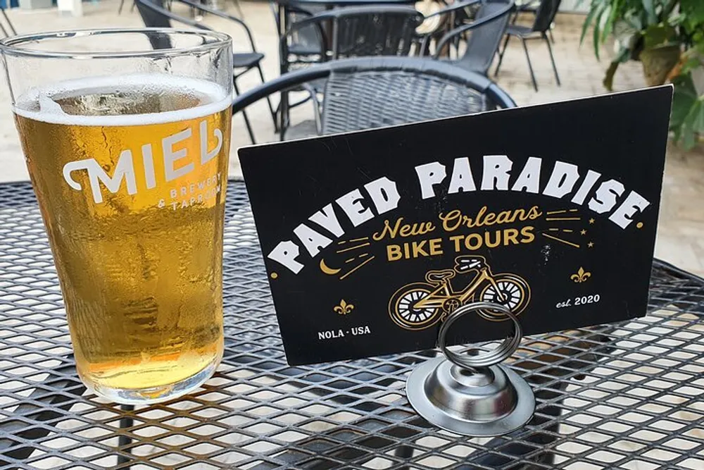 The image shows a glass of beer alongside a promotional table tent for Paved Paradise New Orleans Bike Tours on a metal mesh table indicating a casual outdoor setting possibly at a brewery or a bar