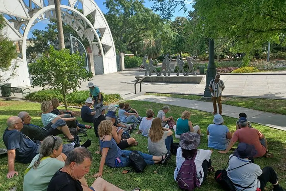 A group of people is sitting on the grass attentively listening to a person standing and speaking in what appears to be a park with a unique arch and sculptures in the background