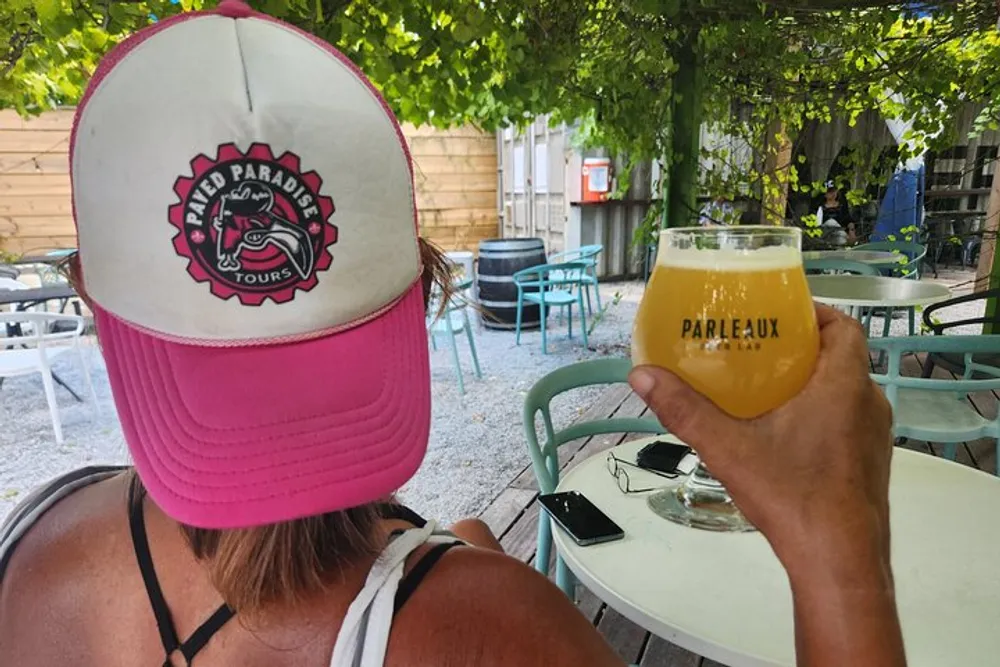 A person is sitting at an outdoor caf wearing a pink and white baseball cap with a logo and holding a glass of beer labeled Parleaux