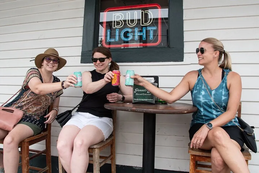 Three women are cheerfully toasting with cans of drink while sitting at an outdoor bar table under a Bud Light neon sign