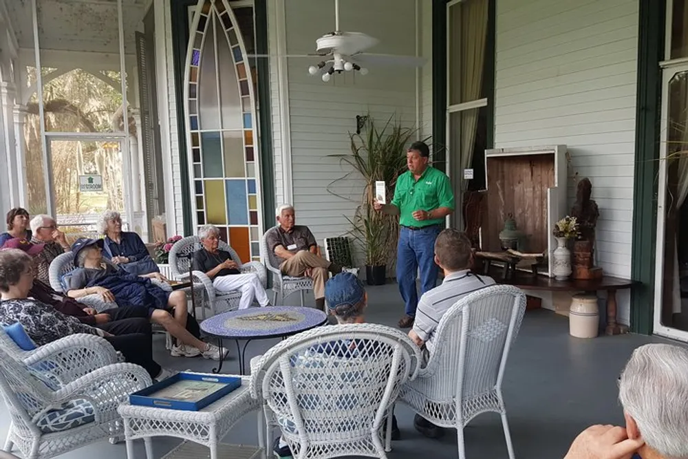 A group of people is attentively listening to a presenter speaking on a porch furnished with wicker chairs and decorated with a stained glass window