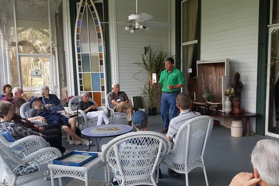 A group of people is attentively listening to a presenter speaking on a porch furnished with wicker chairs and decorated with a stained glass window.