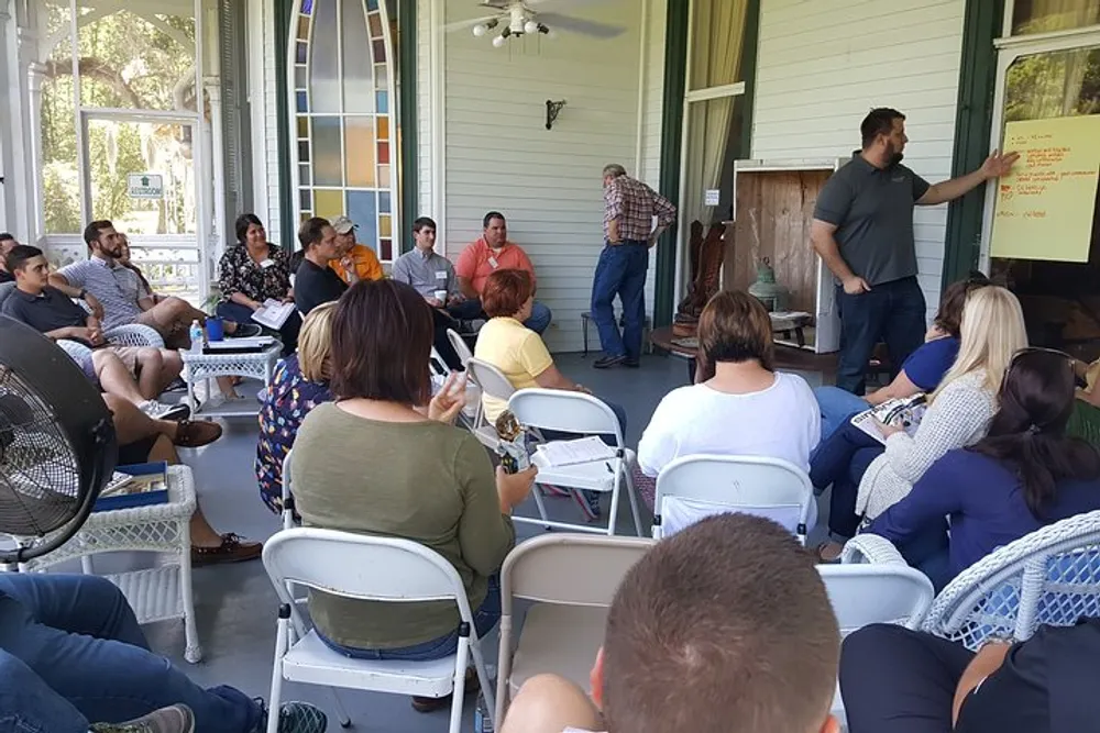 A group of people is attentively listening to a presenter on a porch-like setting with some taking notes and a fan providing comfort from the heat