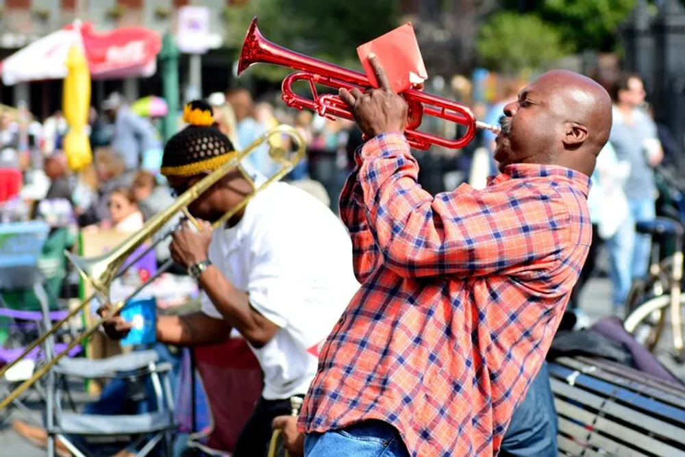 A man passionately plays a red trumpet outdoors with another musician seen playing a trombone in the blurred background among a lively street setting