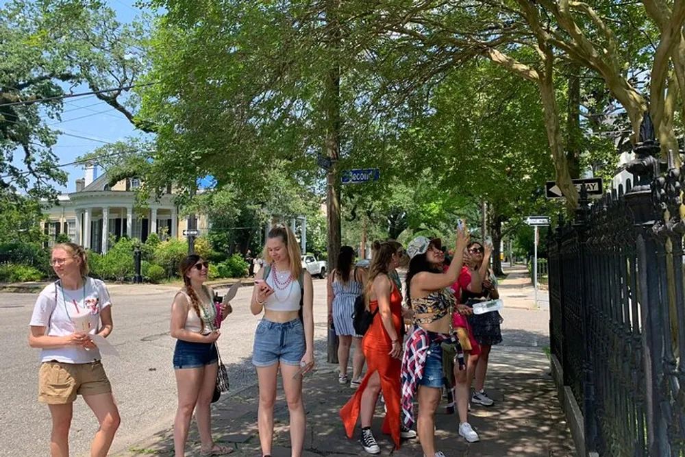 A group of people appears to be enjoying a sunny day outdoors possibly on a sightseeing tour with some individuals engaged in conversation or looking at their phones while standing near a street corner with lush greenery