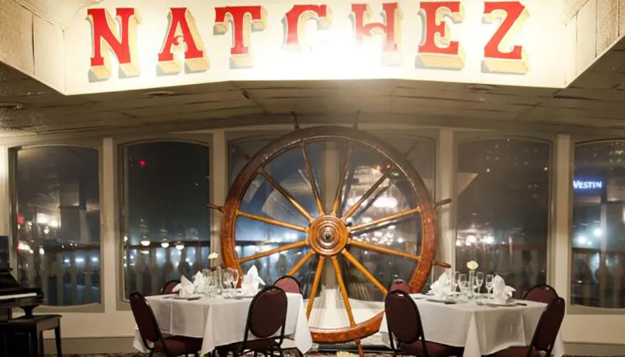 The image shows an elegantly set dining area with a large ship's wheel in the foreground and the word NATCHEZ displayed above in bold letters.