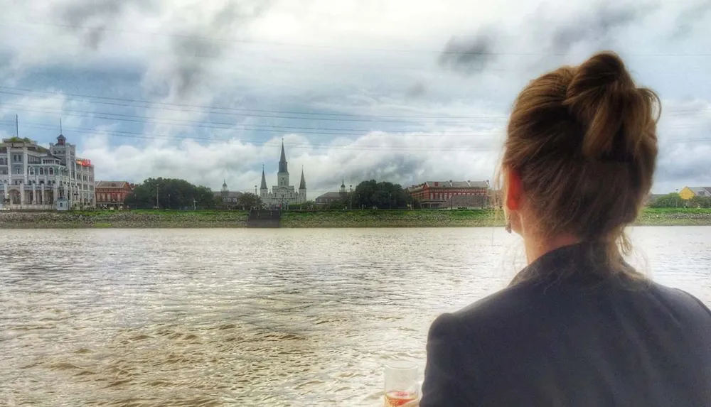 A woman is looking at a riverside view featuring historic architecture under a cloudy sky while holding a drink