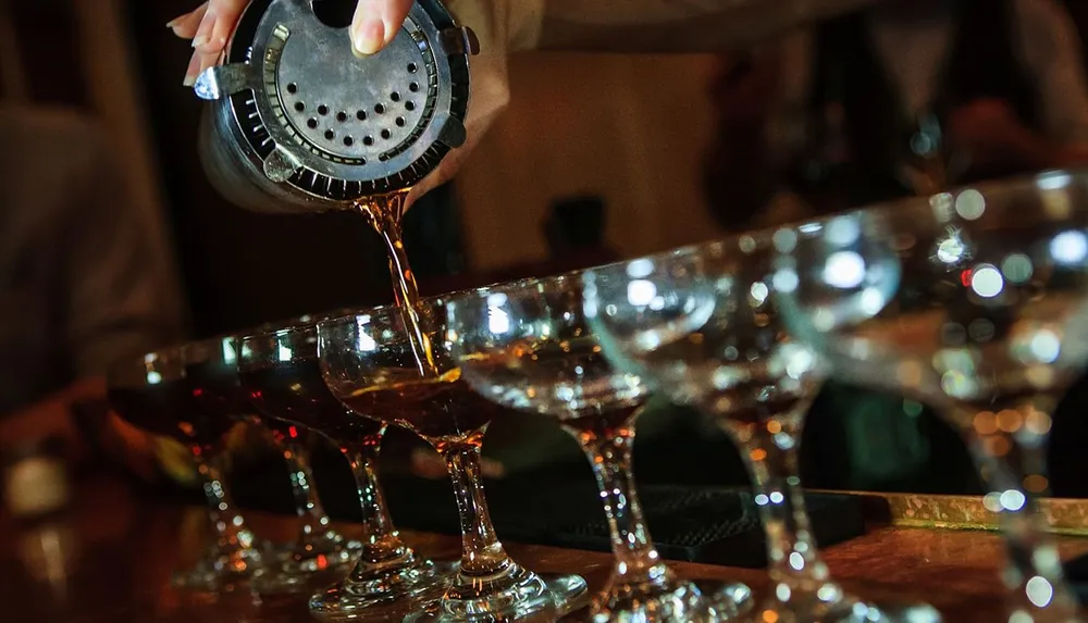 A bartender is pouring a cocktail through a strainer into a row of martini glasses on a bar counter