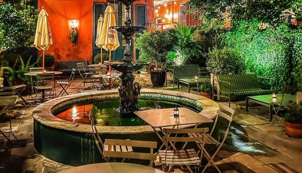 An enchanting evening view of a cozy courtyard with a fountain surrounded by lush greenery and warmly lit walls set up with tables and chairs for dining