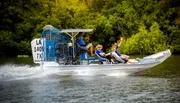 A group of passengers are enjoying a ride on an airboat, skimming over the water with lush green foliage in the background.