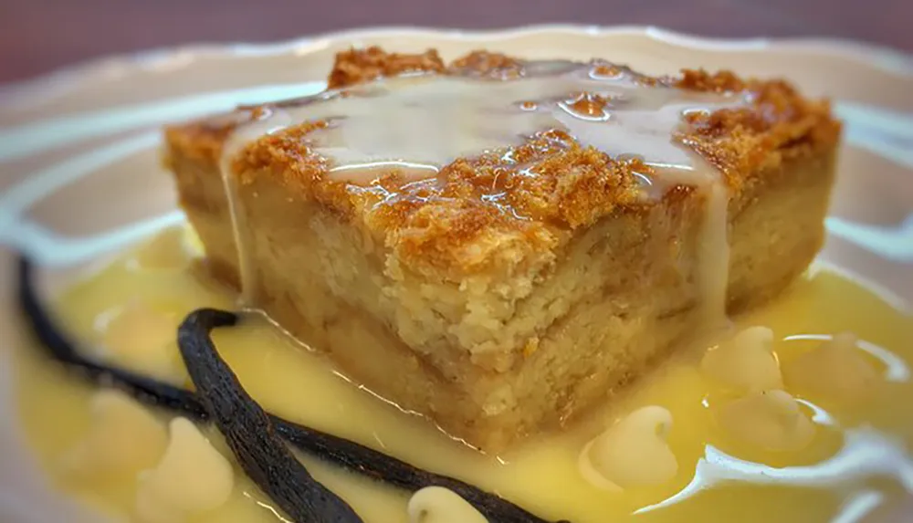 A slice of bread pudding topped with white icing and served with a vanilla bean sauce is presented on a plate
