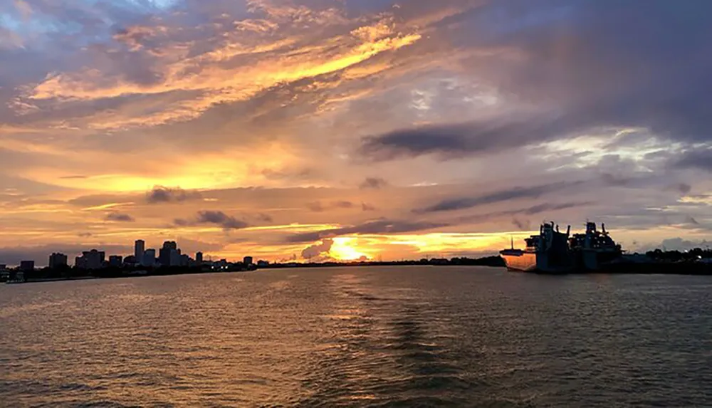 The image captures a vibrant sunset sky with golden and purple hues over a body of water showcasing the silhouette of a city skyline and a large ship to the right