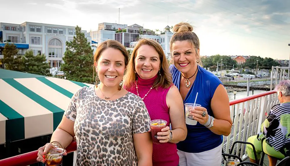 Three women are smiling at the camera while holding beverages with a backdrop of a marina and buildings at what appears to be a rooftop or outdoor gathering