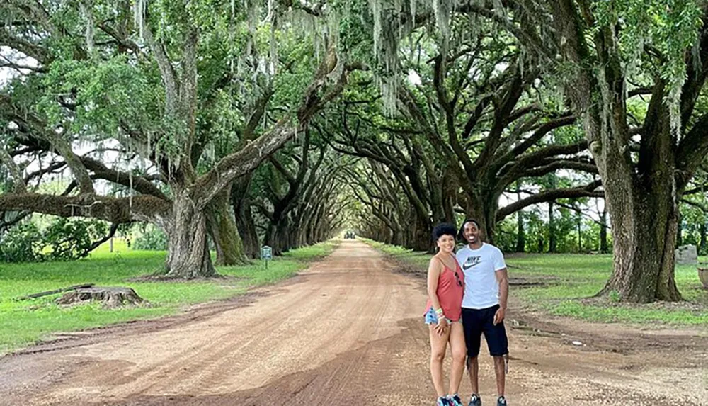 A couple is smiling for a photo on a picturesque tree-lined dirt road with Spanish moss hanging from the oak branches above them