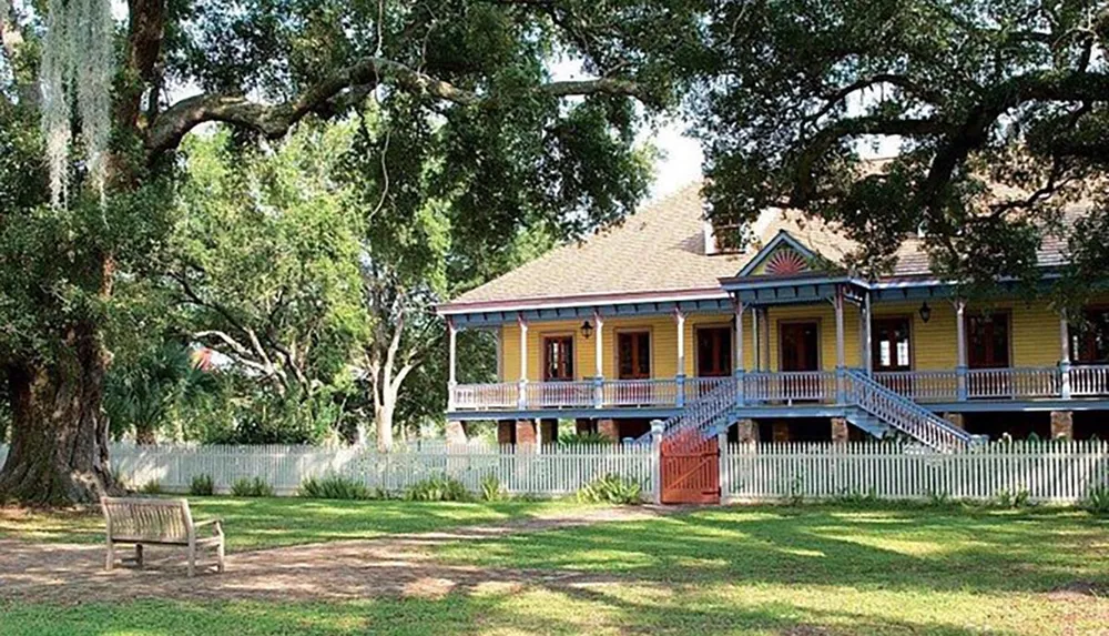 A traditional yellow wooden house with a blue-trimmed porch and a surrounding white picket fence stands amidst lush trees and a lawn with a lone bench