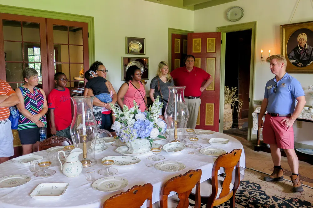 A group of people is attentively listening to a tour guide in a room with antique furniture and a set dining table