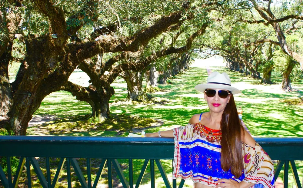 A woman in a colorful blouse and a white hat is posing for a photo with a scenic background of an avenue of sprawling moss-draped trees