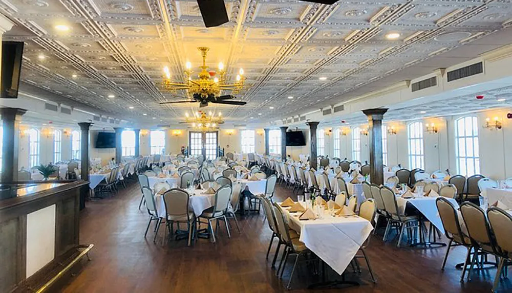 The image shows an elegant banquet hall with decorated tables set for an event featuring a patterned ceiling and chandeliers