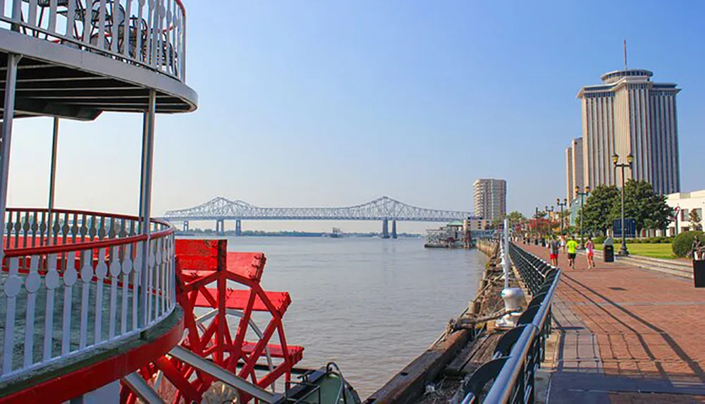 The image features a riverfront with a red paddlewheel a prominent bridge in the background a high-rise building and people walking along a riverside promenade under a clear blue sky