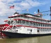 The image shows a classic multi-deck riverboat with a large red paddle wheel named City of New Orleans docked under a clear sky with American flags fluttering atop