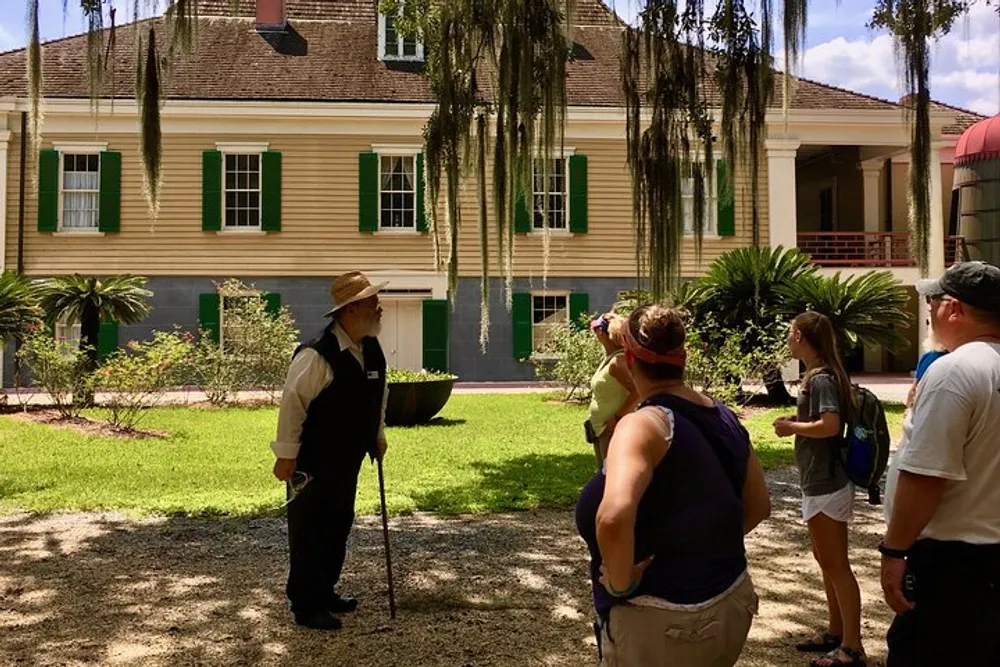 A person in period costume is speaking to a group of visitors outside a large historic house with green shutters and a lush garden setting